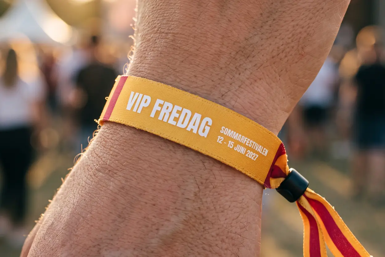 Nahaufnahme eines gelben Stoffbands mit der Aufschrift VIP FREDAG, gestreiften Enden und schwarzem Kunststoffverschluss am Handgelenk